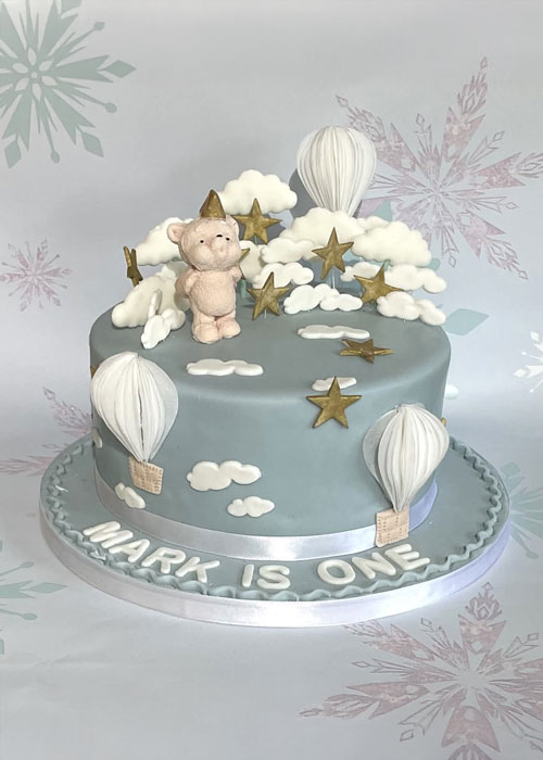 Light blue fondant birthday cake with white clouds, stars, bear and hot air balloons; white icing on board reads "Mark is one"